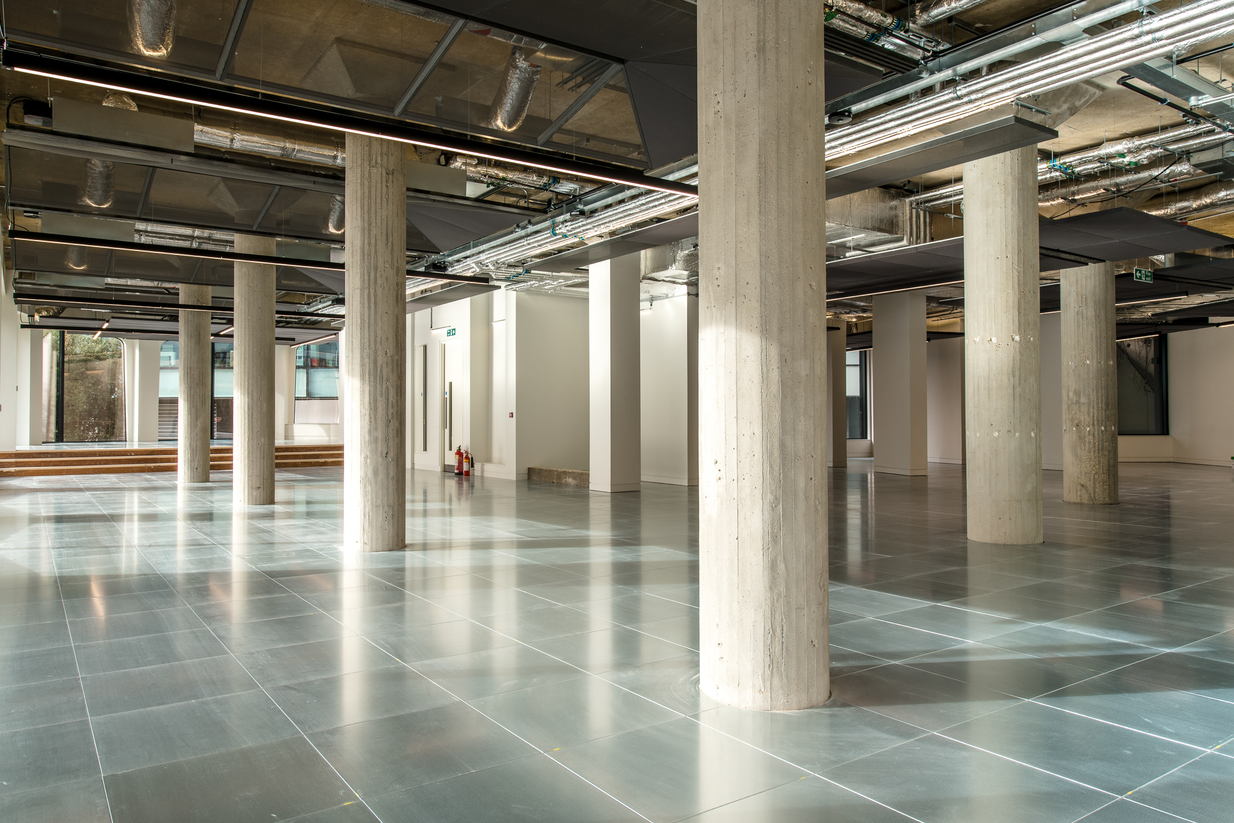 A spacious area with reflective tiled flooring and concrete pillars spread throughout. Large arched windows allow for plenty of natural light, while fire exit signs and modern light fixtures adorn the ceiling.