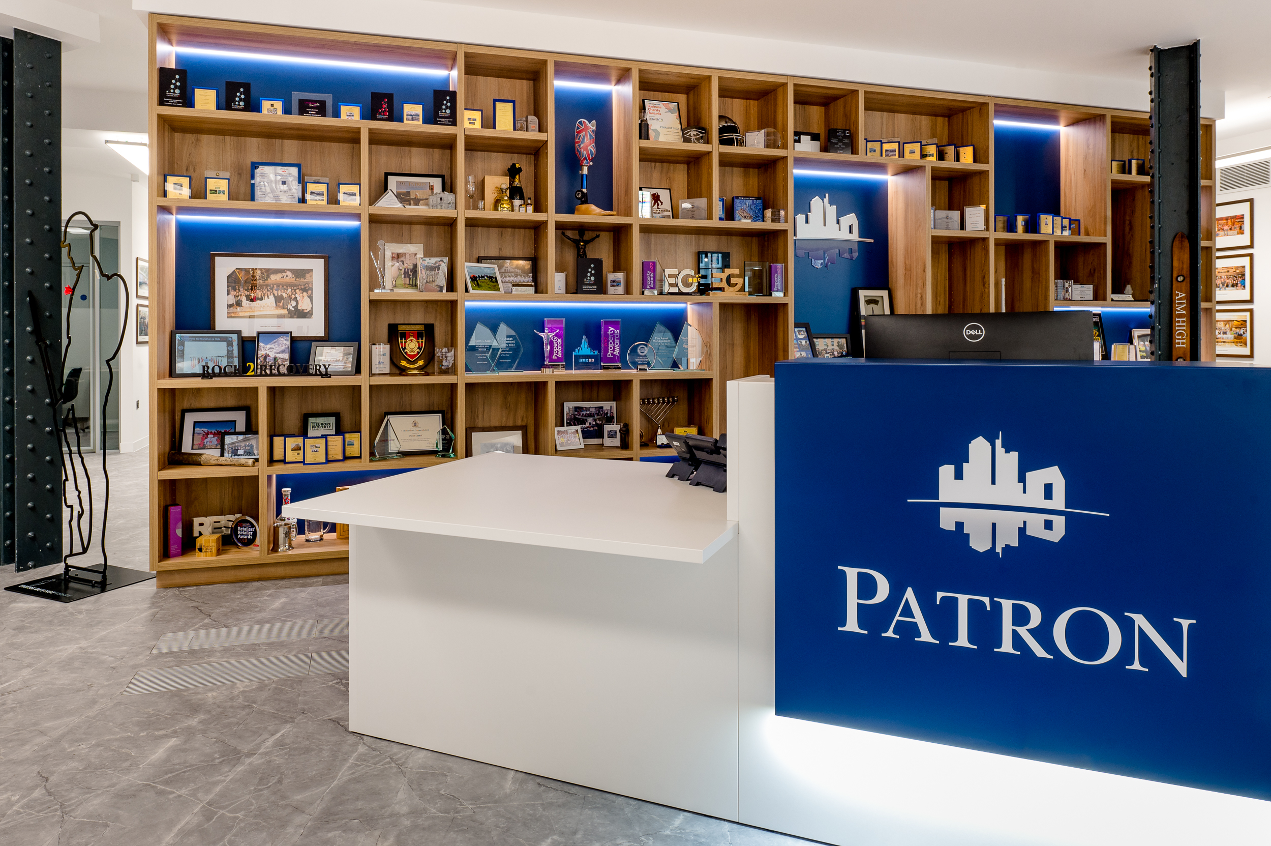 Reception desk with the Patron logo, with a background of wooden shelving displaying various awards.