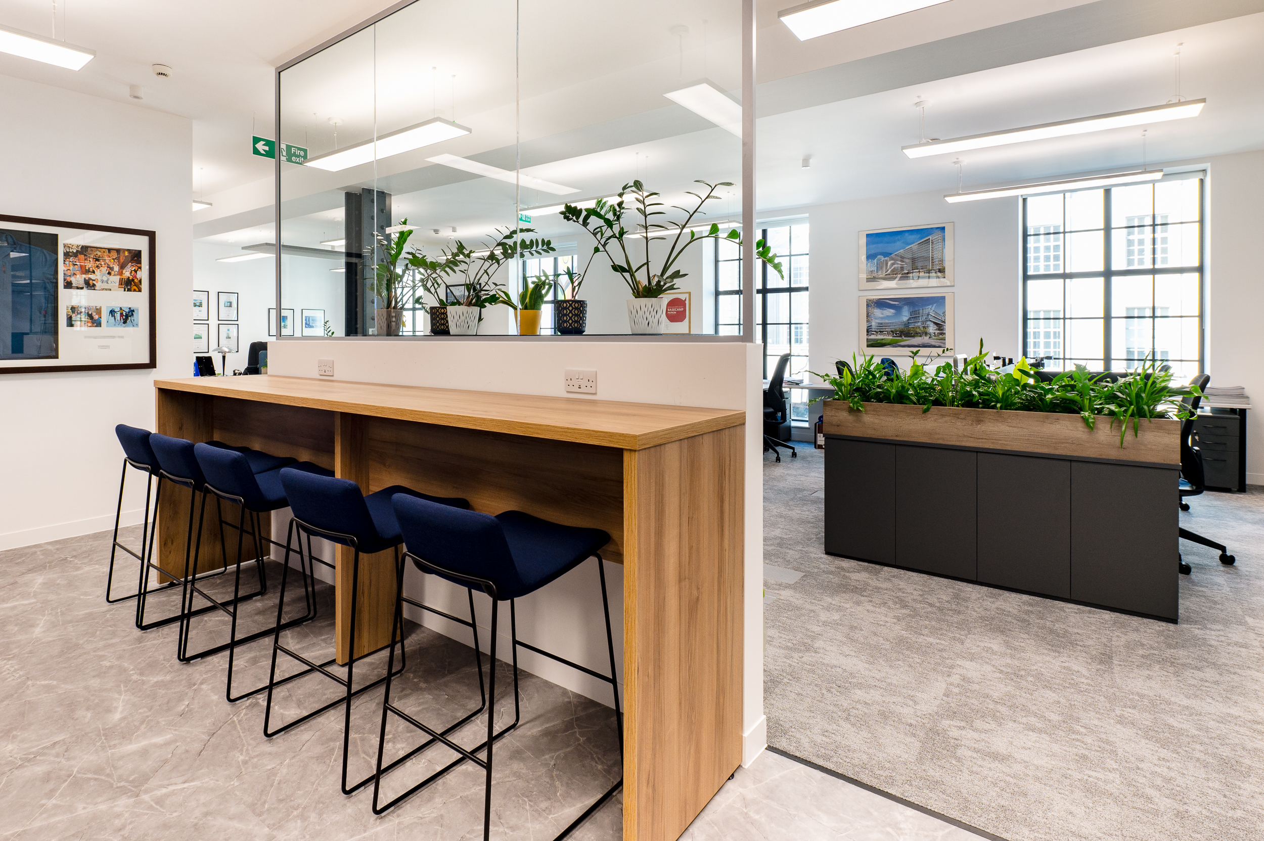 A stylish, casual seating area with a wooden counter and high stools, separated by a glass partition from the main office space.