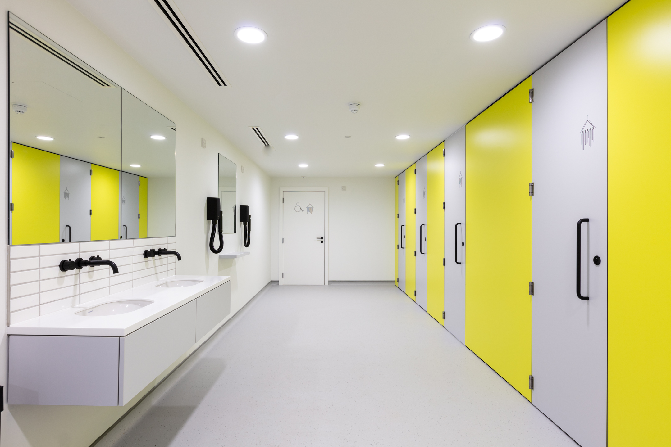A bathroom featuring yellow cubicle doors, white tiled walls, and dual sinks at Witan Gate. The design is bright and welcoming, with modern fixtures.