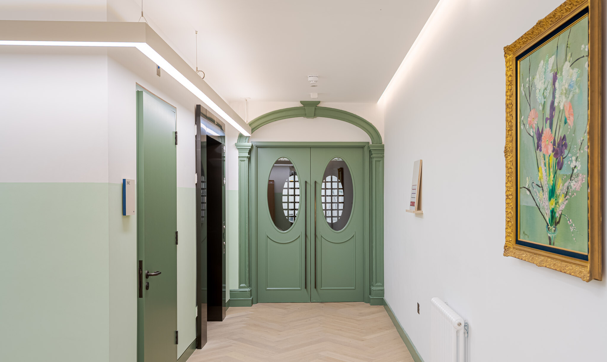 Heritage-style corridor at Bloomsbury House featuring arched double doors in sage green, a soft pastel colour palette, herringbone flooring, and contemporary lighting alongside classic framed artwork.