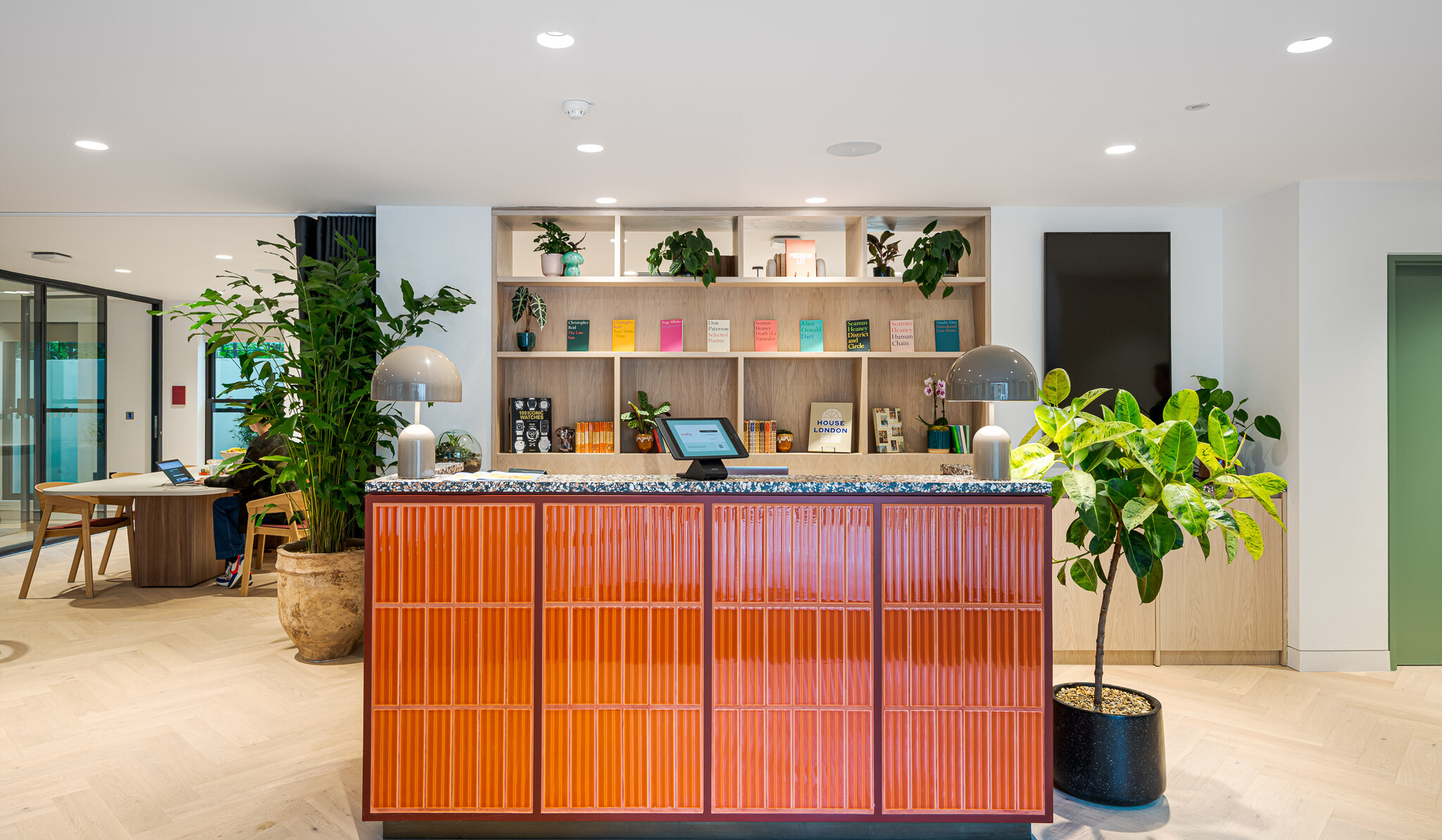 Reception area at Bloomsbury House with a bold orange-tiled counter, terrazzo surface, built-in shelving and curated books, framed by lush indoor planting and soft ambient lighting.