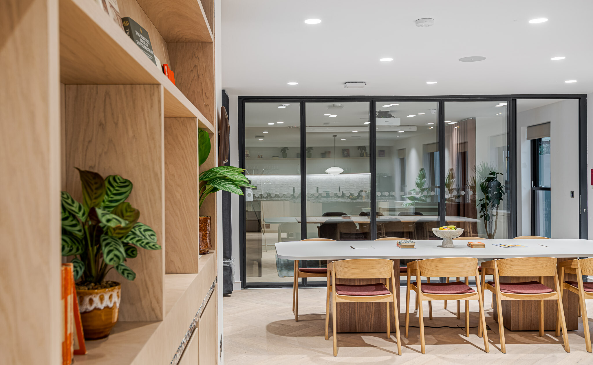 Detail view of communal table and timber shelving at Bloomsbury House, with glass partition revealing a meeting space beyond, styled with indoor plants and soft lighting.