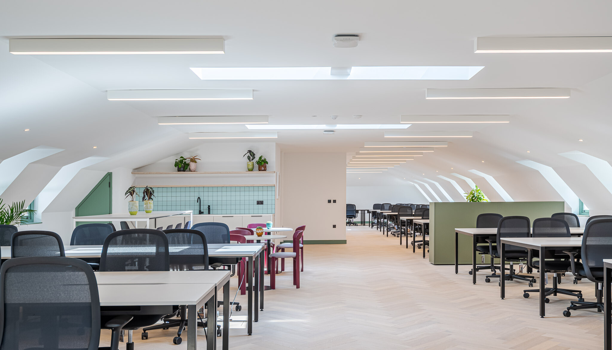 Light-filled attic workspace at Bloomsbury House with open-plan desking, skylights, a breakout kitchenette with green tiling, and splashes of colour from plants and seating.