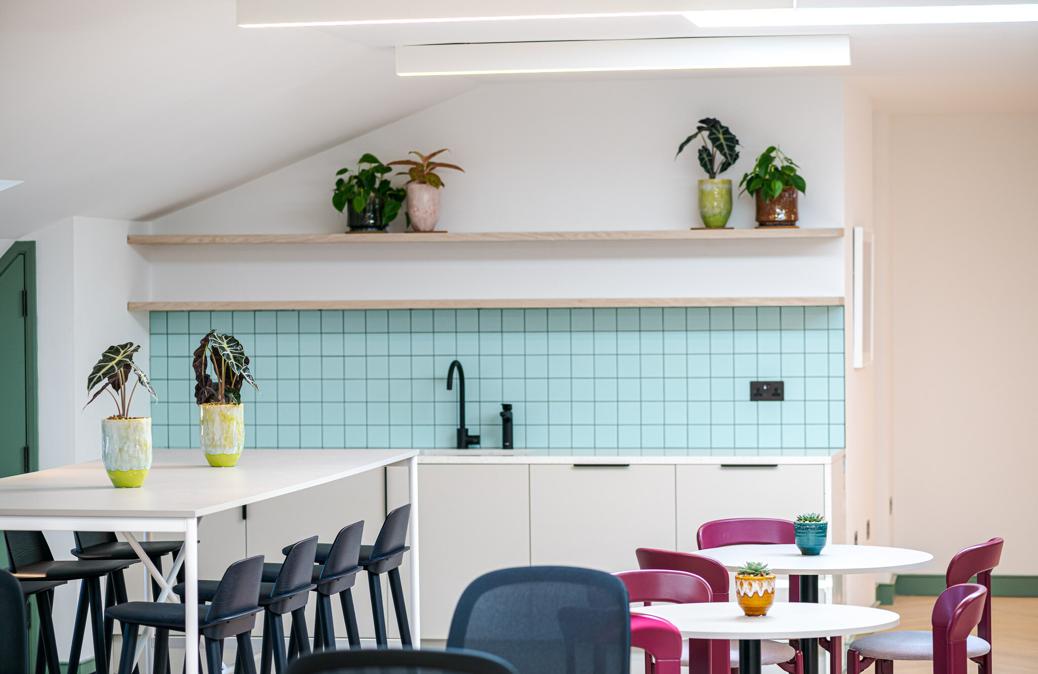 Detail of kitchenette at Bloomsbury House with mint green tiled splashback, matte black tap, and open shelving above, paired with white furniture and colourful potted plants.