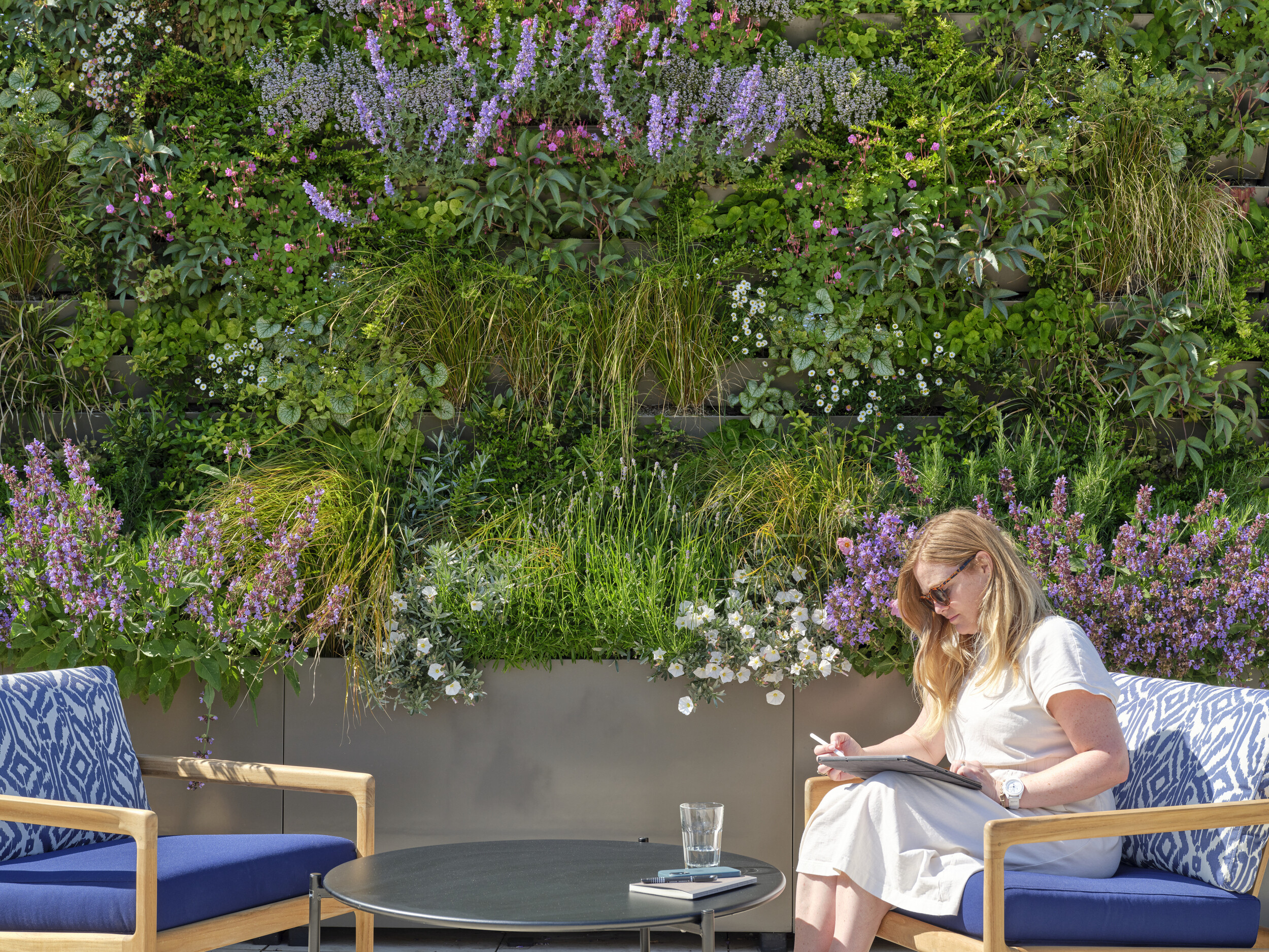 Outdoor seating at The Waterman rooftop by Ambit, with a woman working in front of a lush living wall filled with flowering plants and greenery.
