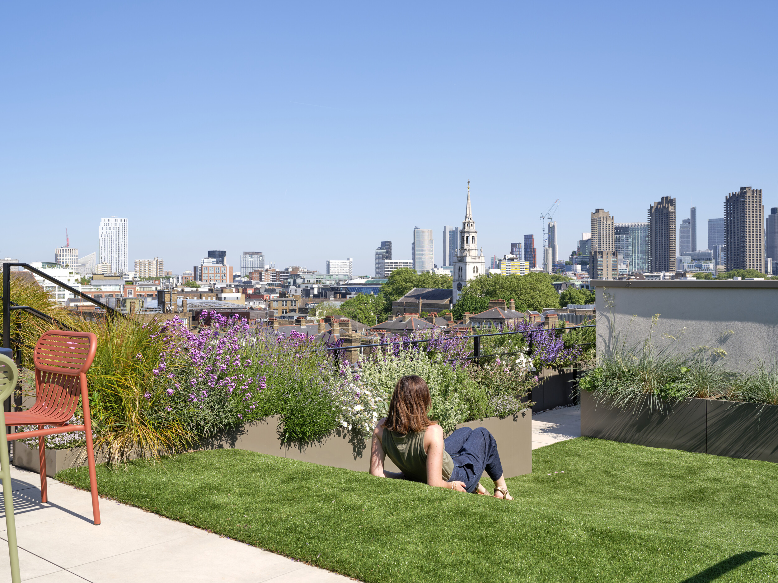 Panoramic view from The Waterman rooftop by Ambit, with wildflower planting and lawn seating overlooking the London skyline under a clear blue sky.