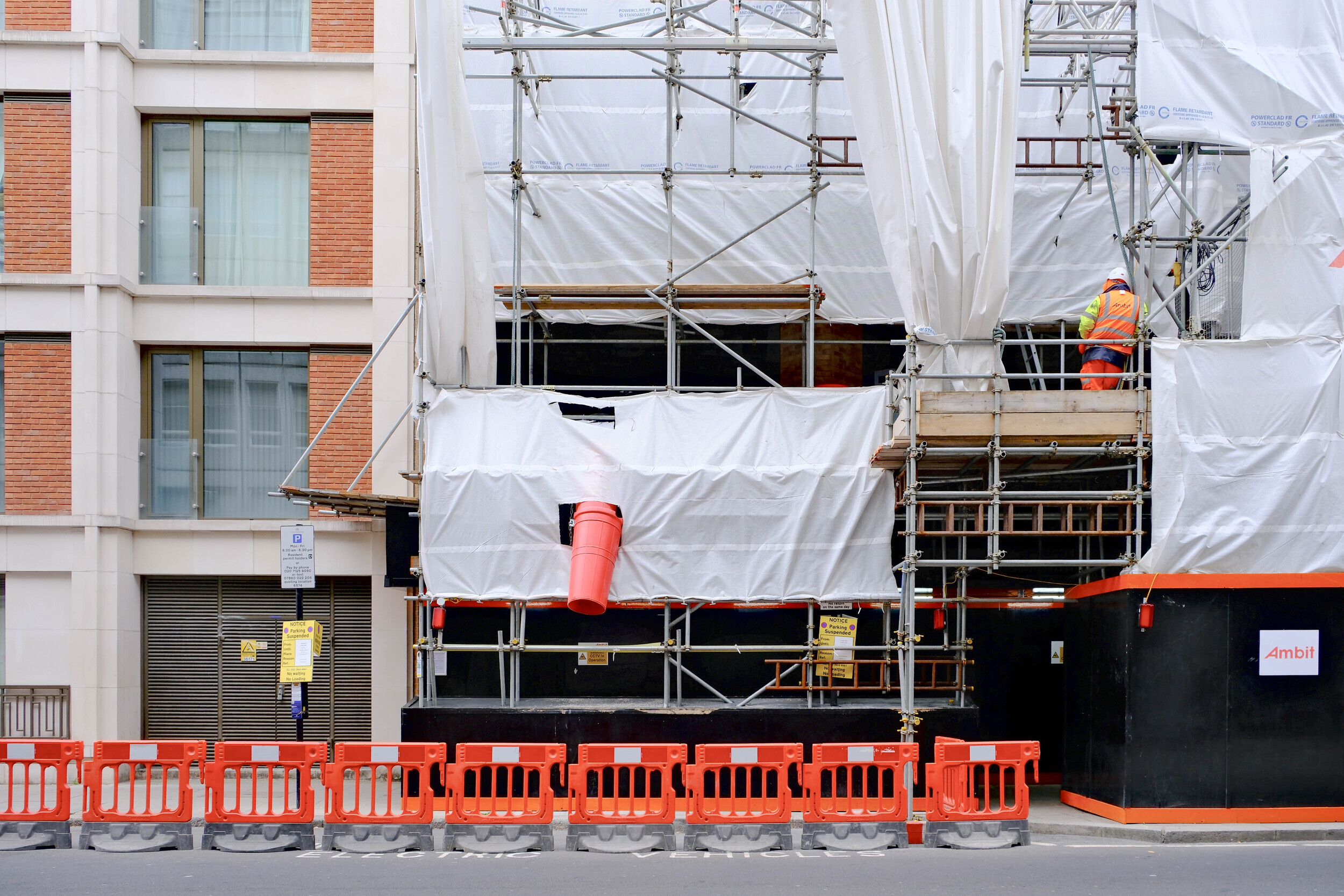 Scaffolding-covered building façade with white sheeting, a red waste chute, and orange barriers along the pavement, highlighting progress in the Rochester Row Recycling project.