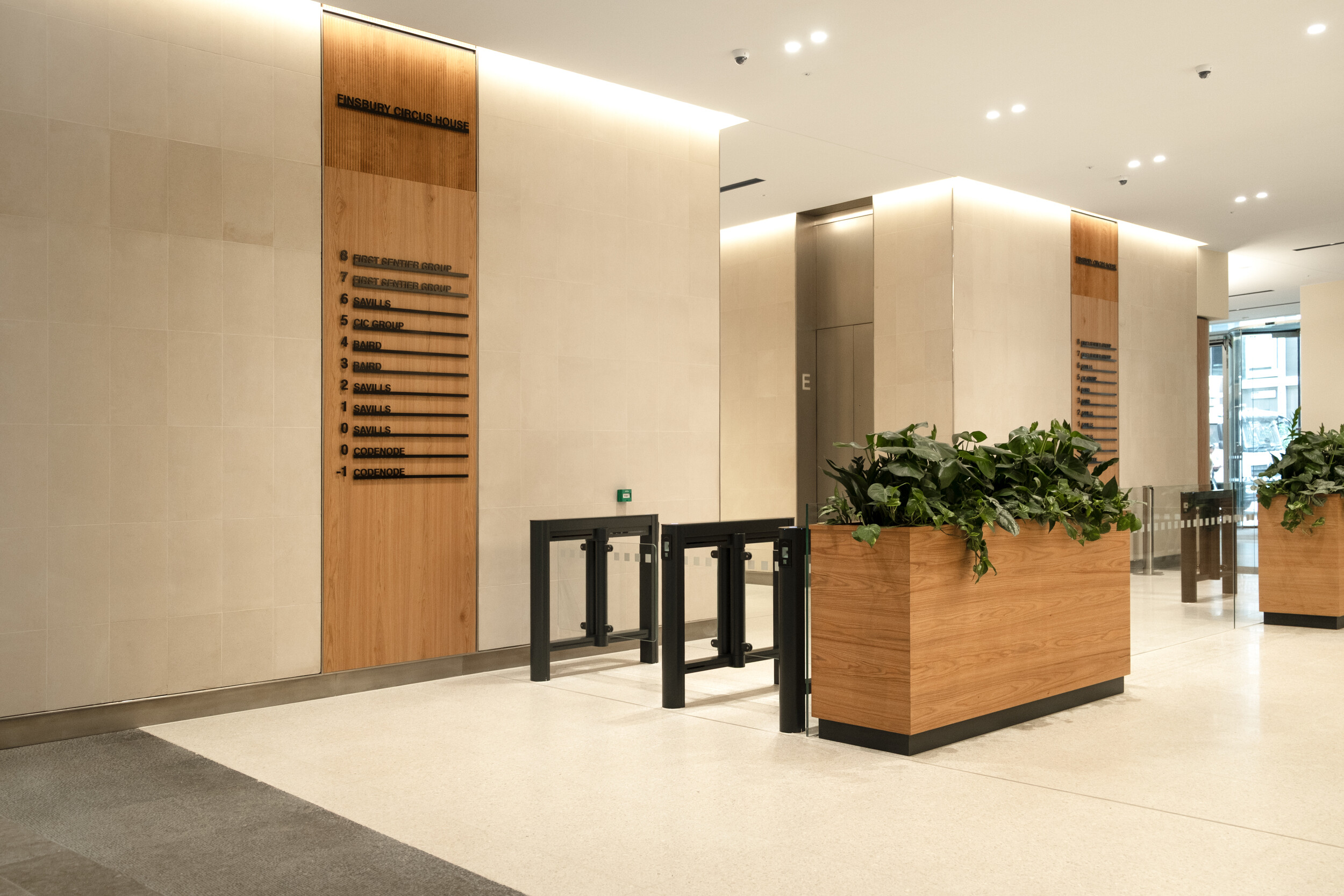 Lift lobby and security gates at Finsbury Circus House, featuring timber wall panelling, illuminated tenant signage, and contemporary planters.