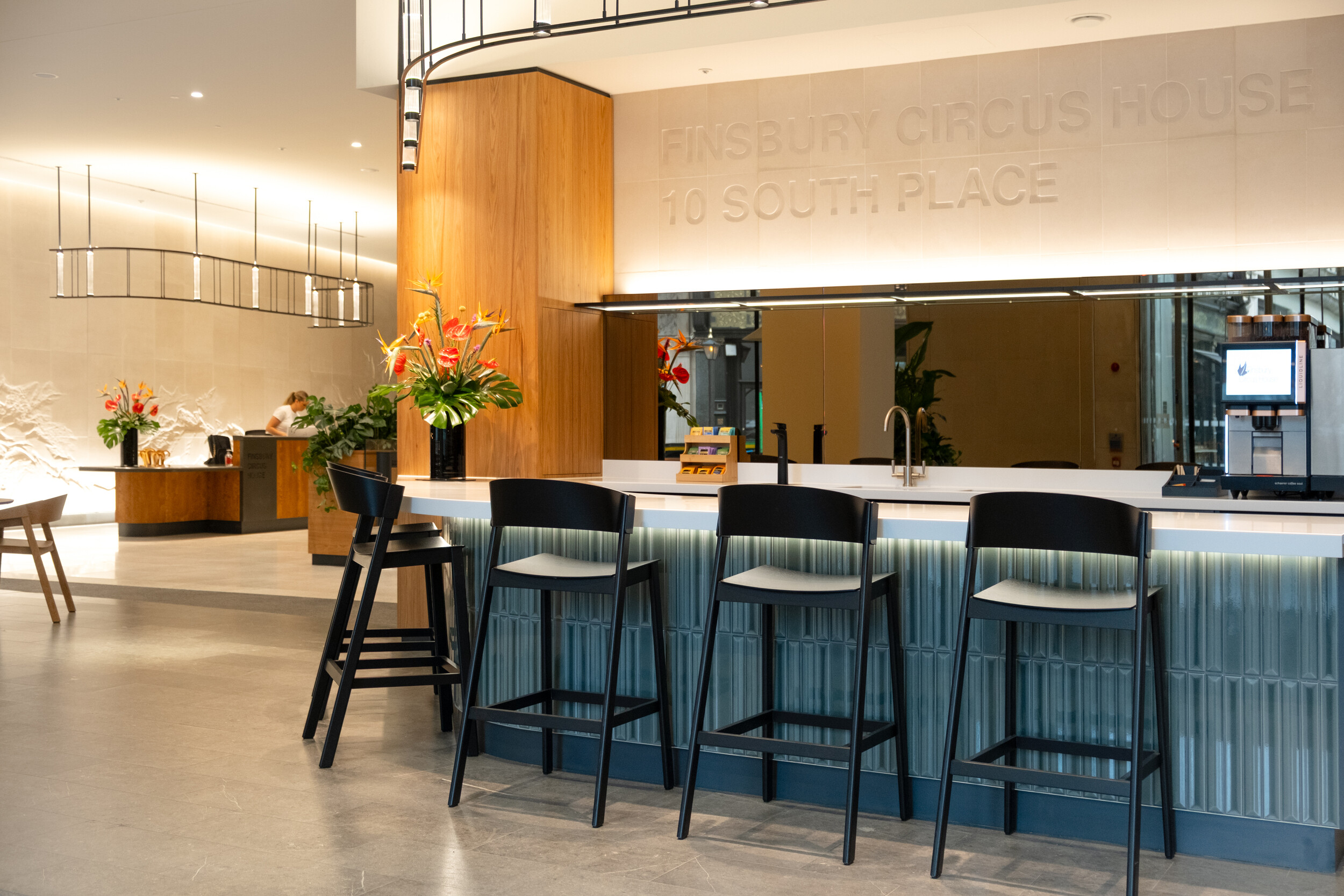 Welcome bar and teapoint at Finsbury Circus House with fluted panelling, black stools, and bold floral arrangements, positioned beneath engraved wall signage as part of the refurbished reception.